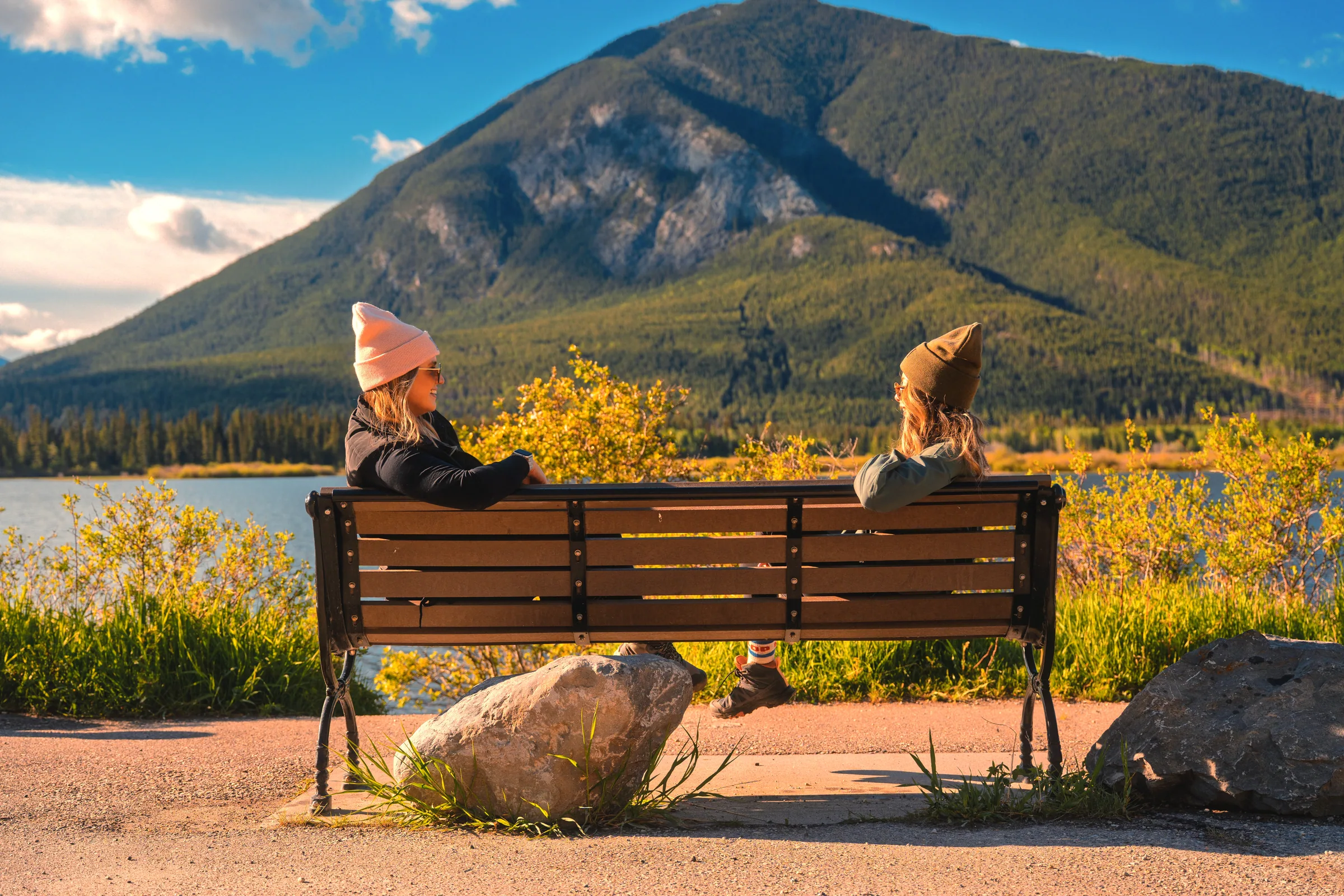 Two visitors resting on a bench beside a paved lakeside path and mountain view.